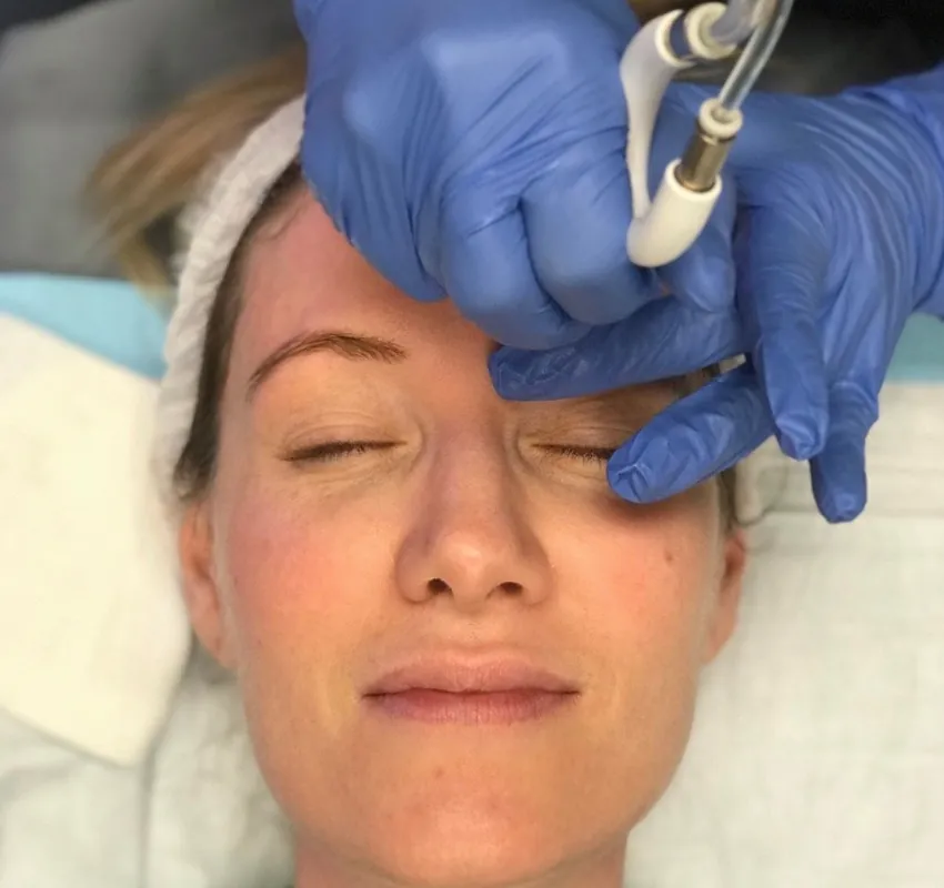 A woman undergoing a facial treatment with a skincare device applied to her forehead by a professional wearing blue gloves.