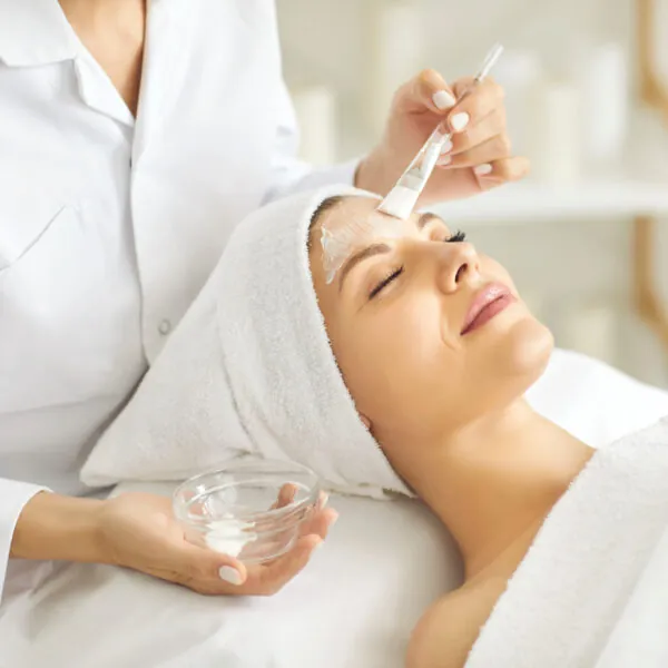 Beautician doing facial treatment and applying beauty mask on face of young woman Beautician in medical coat applies facial mask on woman's face for fresh glowing complexion. Young girl under white towels enjoying skin care treatment at beauty parlor, luxury resort or spa center